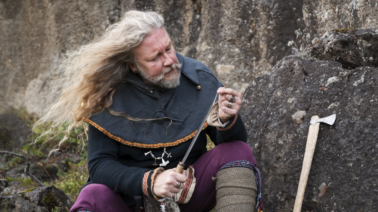 Blond Viking male reenactor in full warrior armour and battle gear is sitting on a rock cleaning his knife in the historic location where Vikings assembled annually to recite and discuss laws, Thingvellir National Park, Pingvellir, Iceland